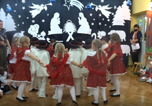 Na tle bożonarodzeniowej dekoracji tańczą dzieci w strojach góralskich w dwóch kółeczkach: chłopcy w środku, dziewczynki na zewnątrz.